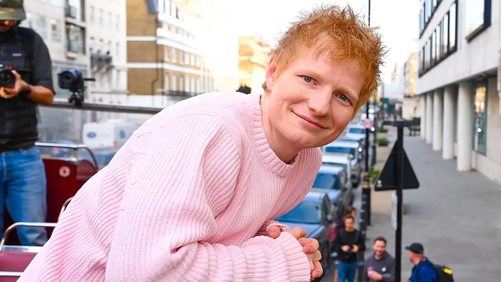 Ed Sheeran met rossig haar in een roze trui, glimlachend op straat met camera's op de achtergrond